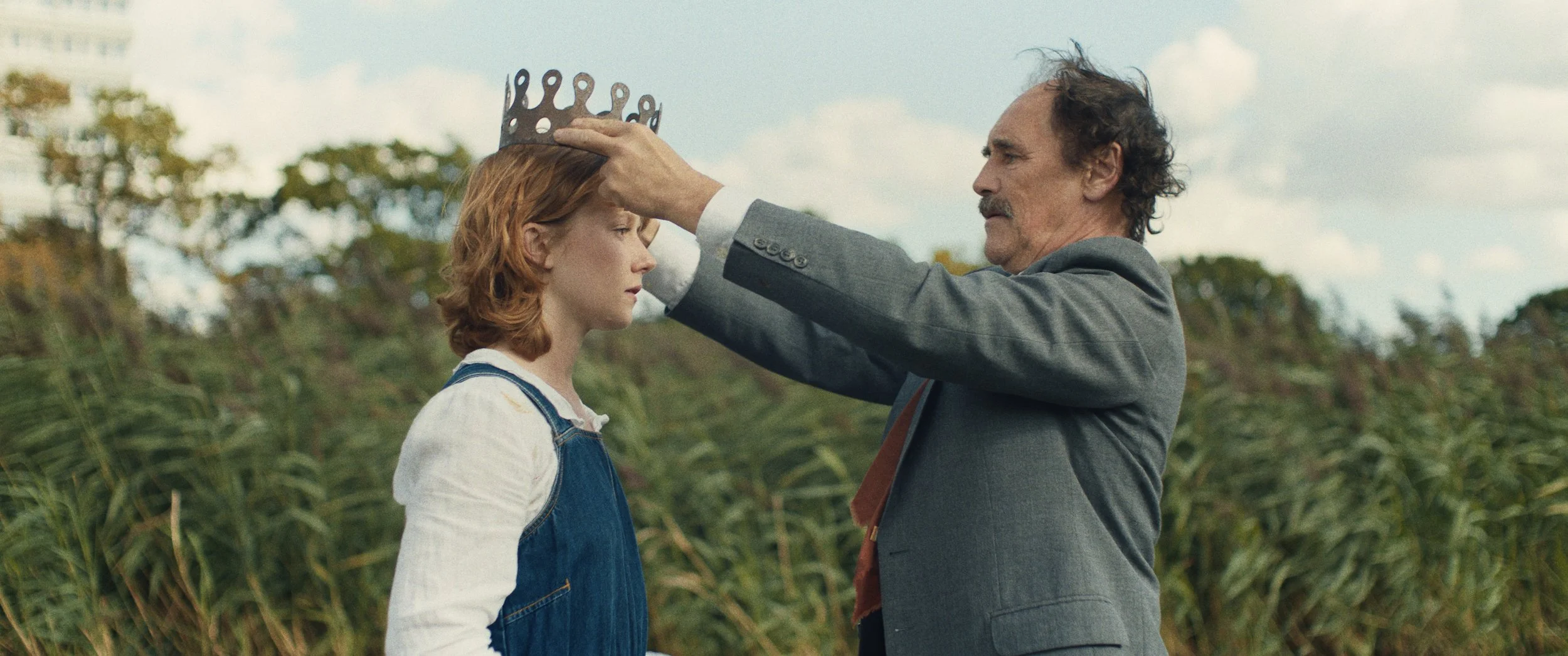 A still from the film Spirit of Place, with actor Mark Rylance wearing a gray suit placing a crown on Hannah Chinn's head with a backdrop of the greenery of the wetlands, with a cloudy sky overhead.