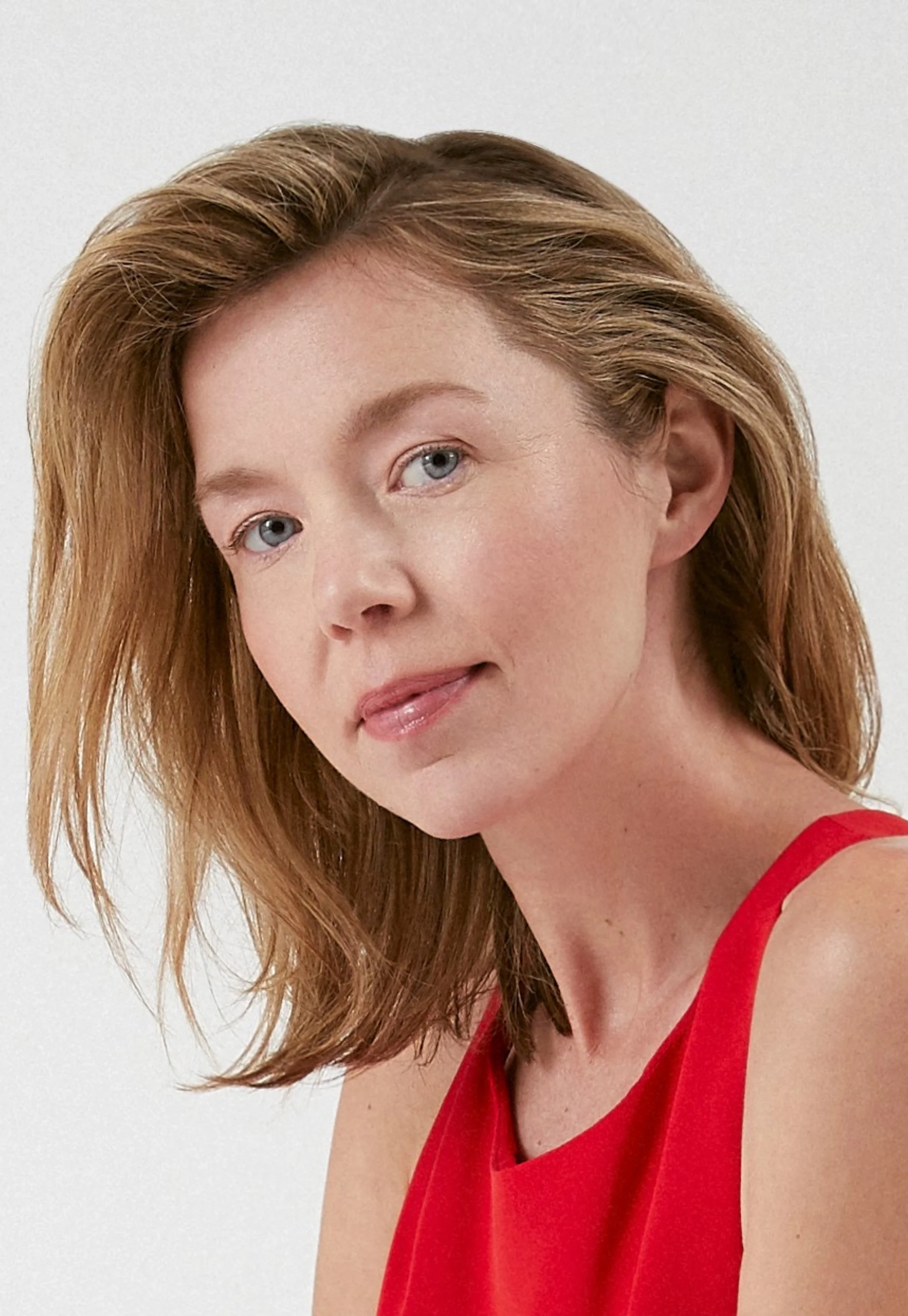 Headshot of Anna Maxwell-Martin with shoulder-length blonde hair wearing a red sleeveless top, smiling softly, against a plain white background.