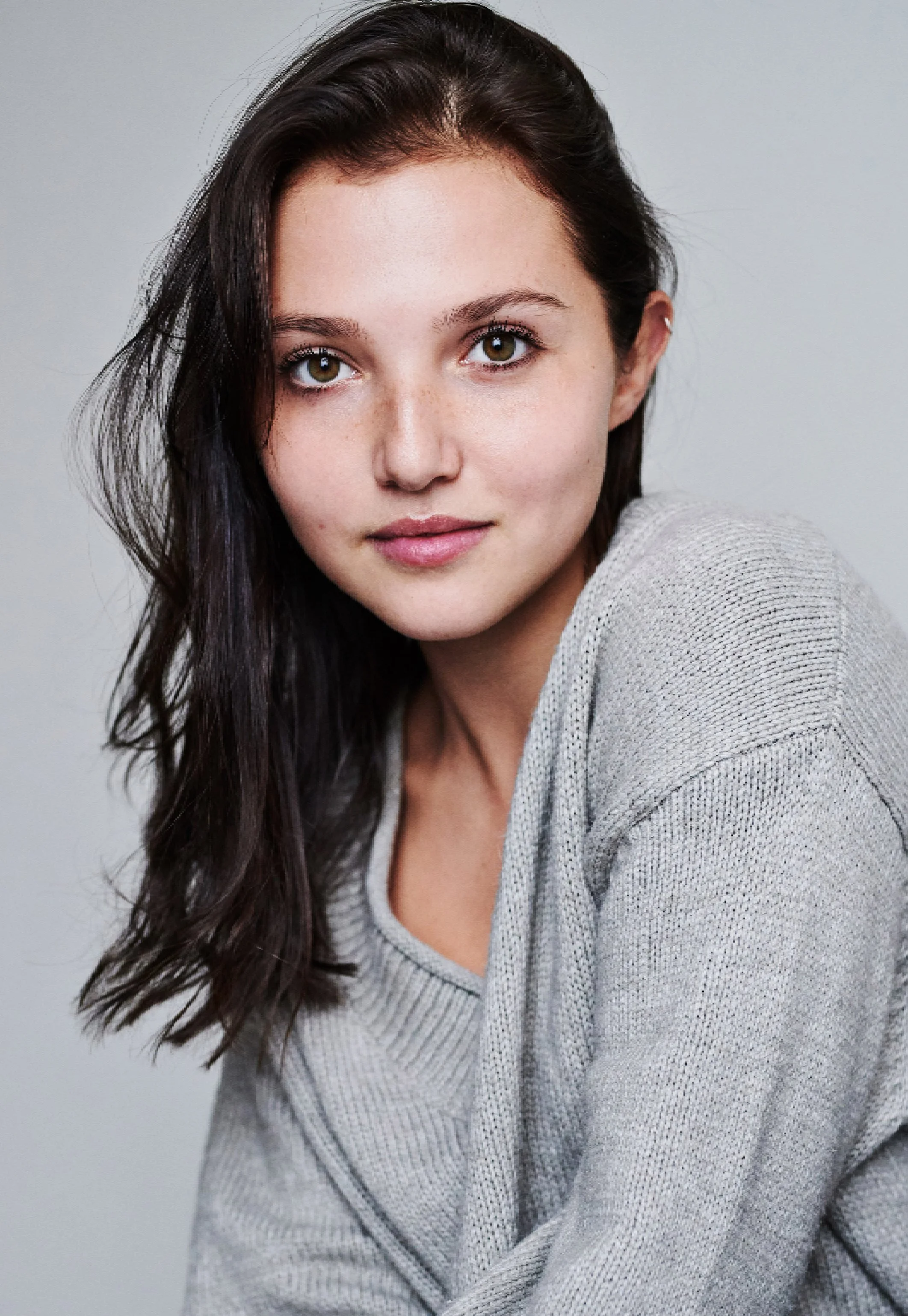 Headshot of Mia McKenna-Bruce with long dark hair, wearing a light gray sweater, looking at the camera against a plain light background.