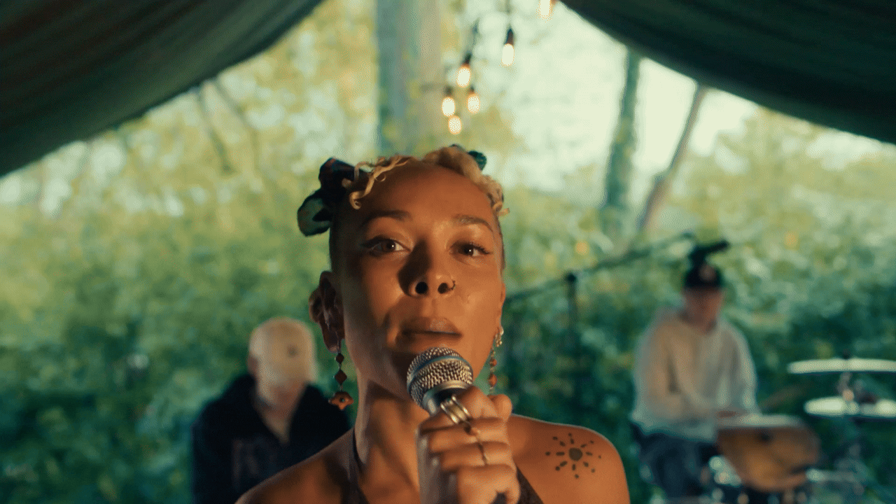 A gif of musician Iyamah singing into a microphone at an outdoor gig, with musicians Kam-BU and Oma playing in the background with lush greenery surrounding the scene.