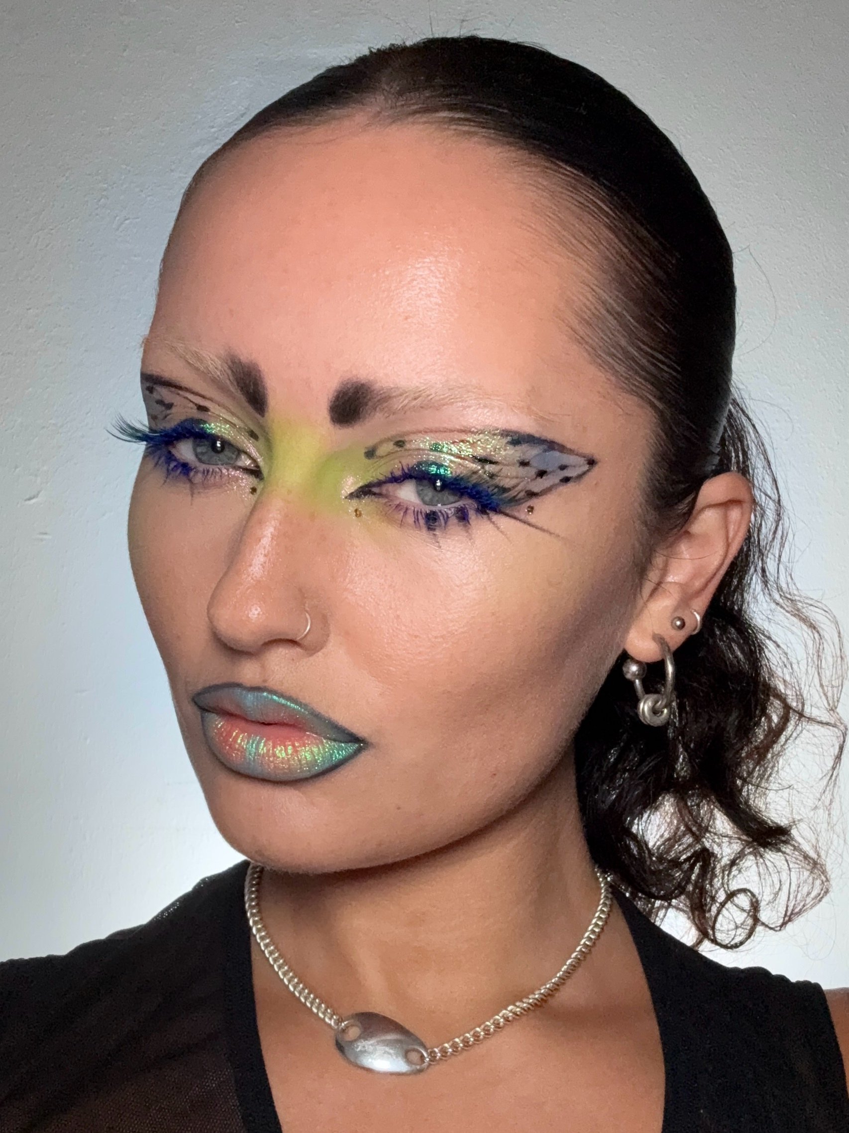 Frankie Darling with a makeup look inspired by the cicada. With lips painted in blues, greens and pinks, and eye makeup green over the nose, blue lashes and black spotted details across he lids, with iridescent greens and blues.
