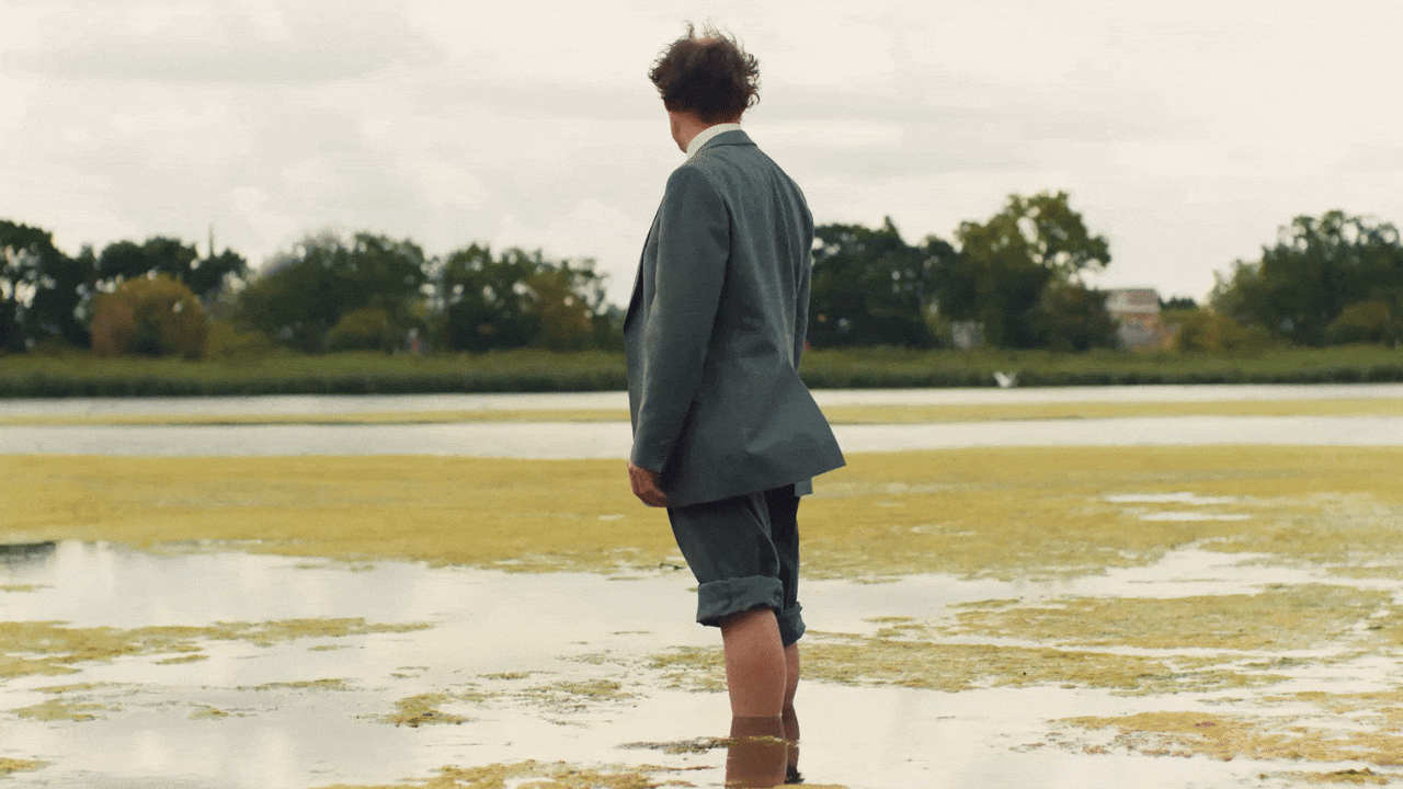 A gif from the film Spirit of Place, with actor Mark Rylance standing side on in the wetlands wearing a gray suit with the trousers rolled above the knees, with a shirt and red tie, looking away from camera and then turning towards camera.