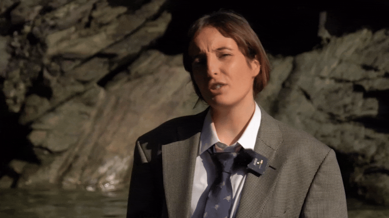 Gif of comedian Spaghetti Sean with tied back brown hair, wearing an oversized blazer, white shirt, and loose dark tie, standing in the sea in front of some rocks talking to camera.