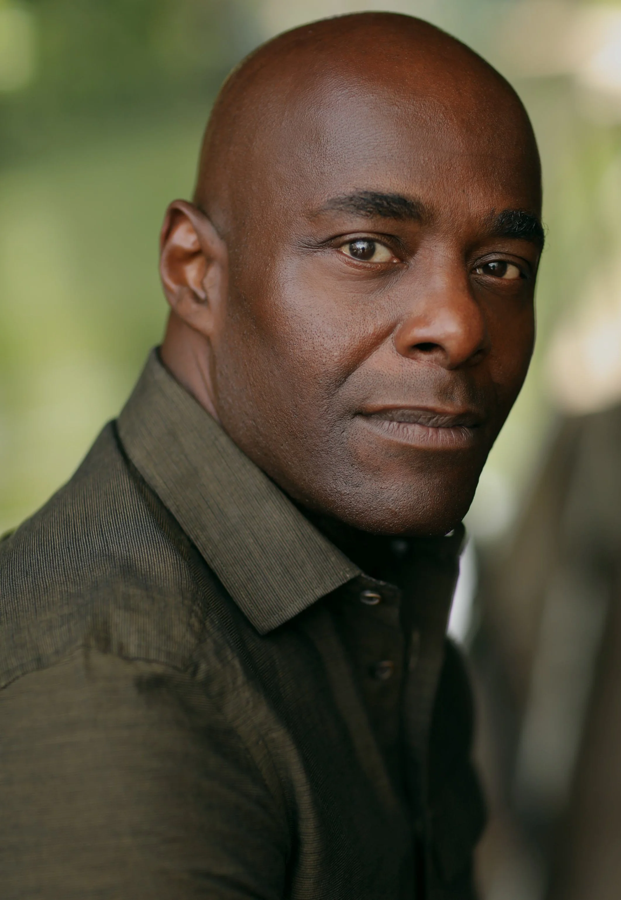 Headshot of Paterson Joseph wearing a dark shirt, looking at the camera with a soft smile against a blurred green background.
