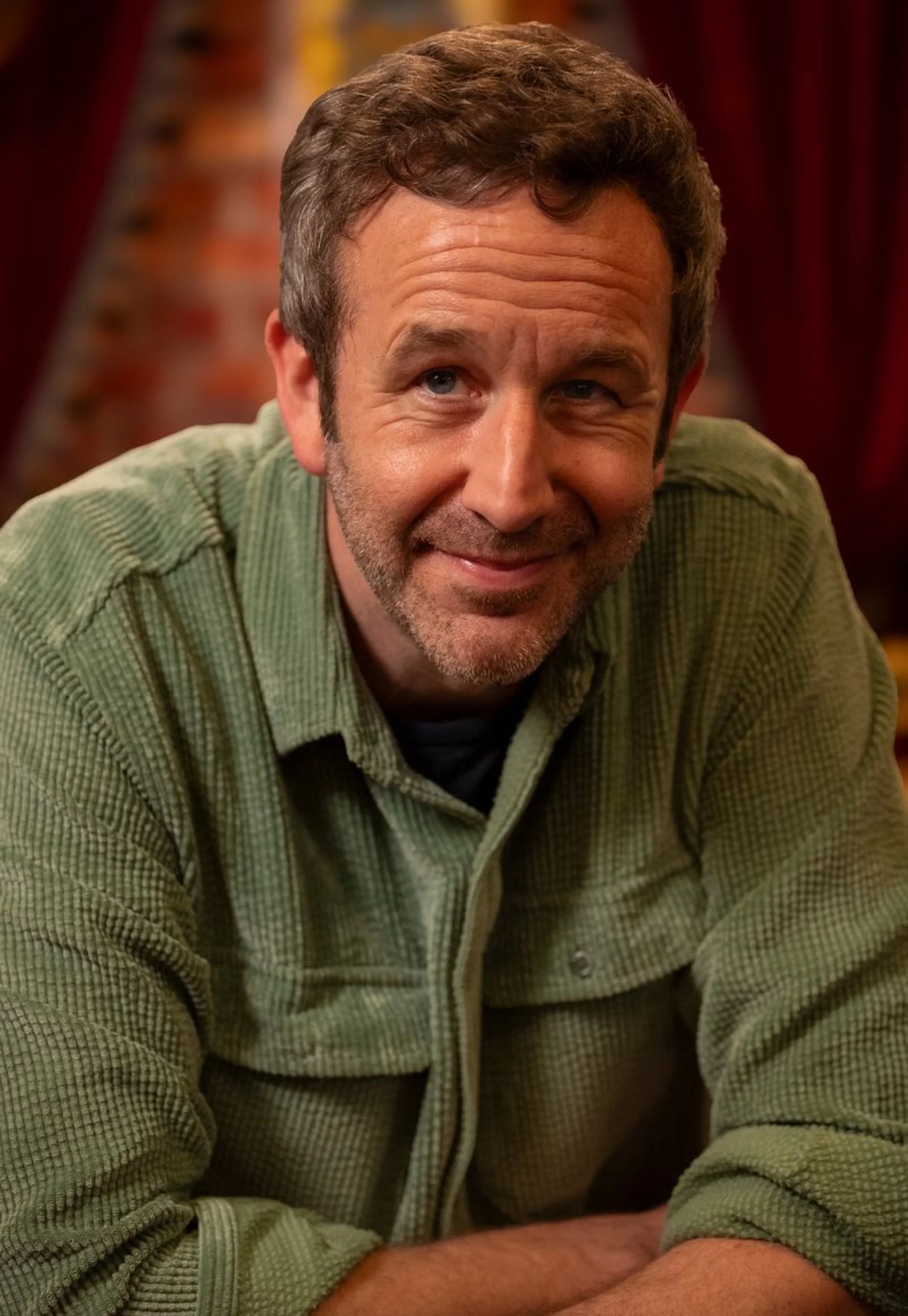 Headshot of Chris O'Dowd with short brown hair and a beard, wearing a green plaid shirt, smiling and leaning forward in a cozy indoor setting with warm lighting and blurred background.