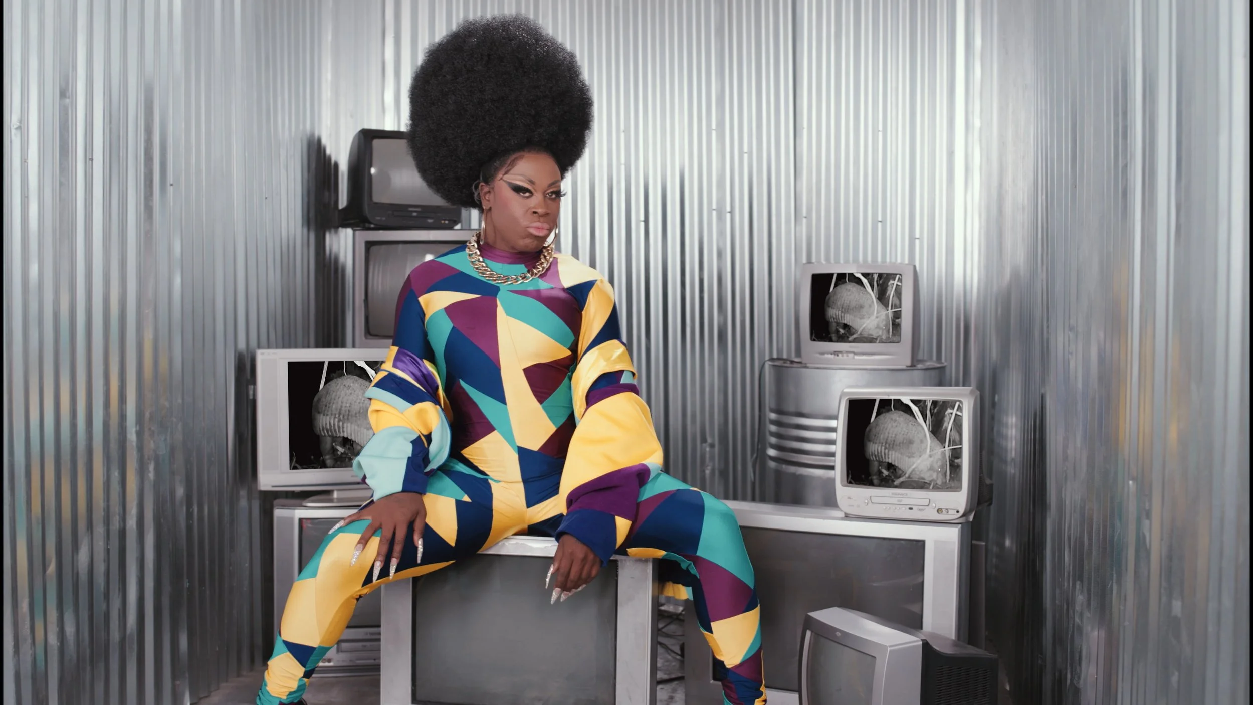 Bob The Drag Queen wearing a colourful patterned outfit with large earrings, a chunky chain necklace, sitting in a room with metallic corrugated walls surrounded by old televisions, some of which have camera trap footage of the Giant Armadillo on.