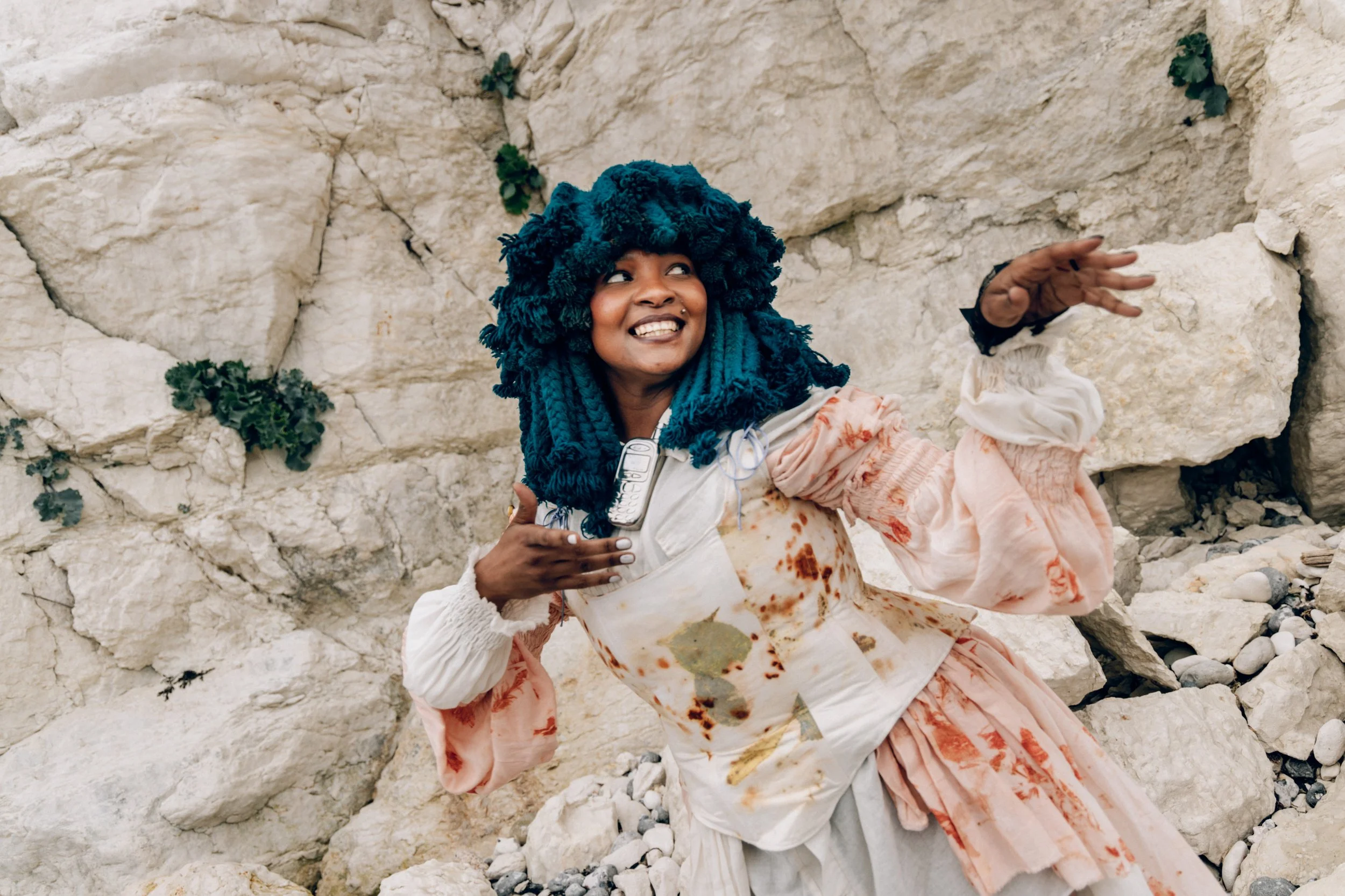 Musician Moonchild Sanelly posing against a rocky background, with her blue braided hair, wearing a floral patterned dress with long, flowy sleeves, smiling and holding a recording device in one hand.