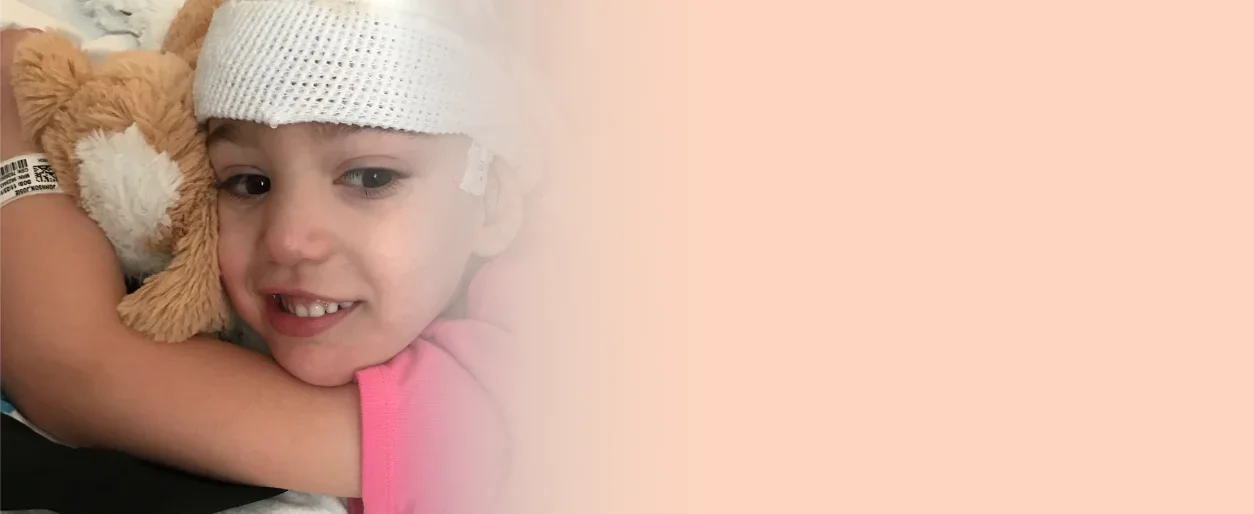 Young girl in a hospital bed with a bandaged head, smiling and holding a stuffed animal.