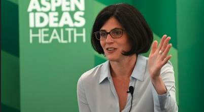 Woman with dark hair and glasses speaking at Aspen Ideas: Health event, green background.