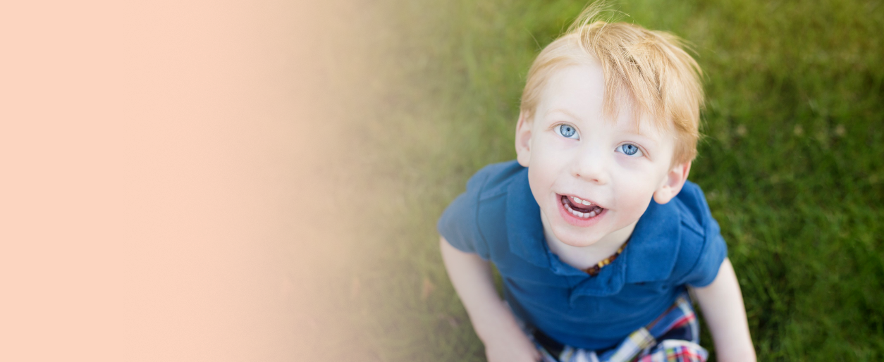 A young boy with red hair and blue eyes looking up and smiling outdoors on grass.