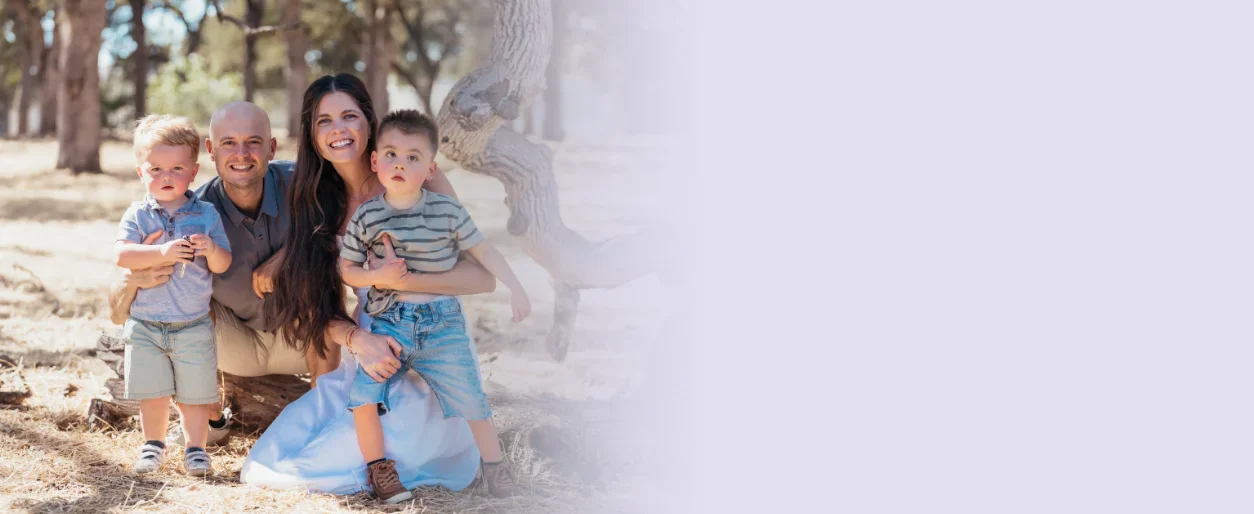 A happy family of four outdoors in a wooded area, sitting on a log, smiling at the camera. The family includes two young children, a father, and a mother.