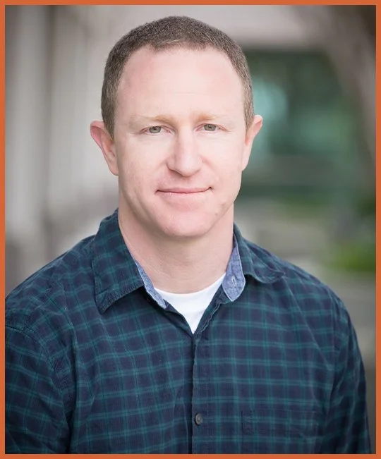 Portrait of a man with short hair, wearing a green and blue plaid shirt, standing outdoors with a blurred background.