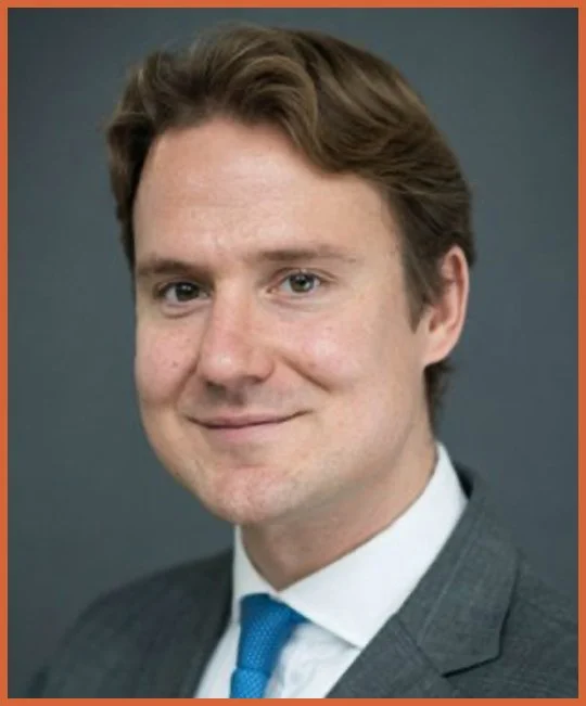 A portrait of a young man with light skin, brown hair, and blue eyes wearing a gray suit, white shirt, and blue tie, smiling slightly against a gray background.