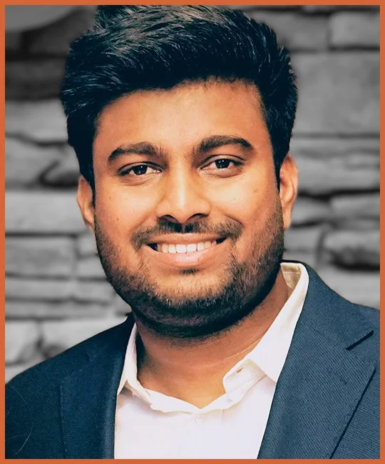 Portrait of a smiling man with dark hair and beard, wearing a dark suit and white shirt, in front of a textured stone wall.