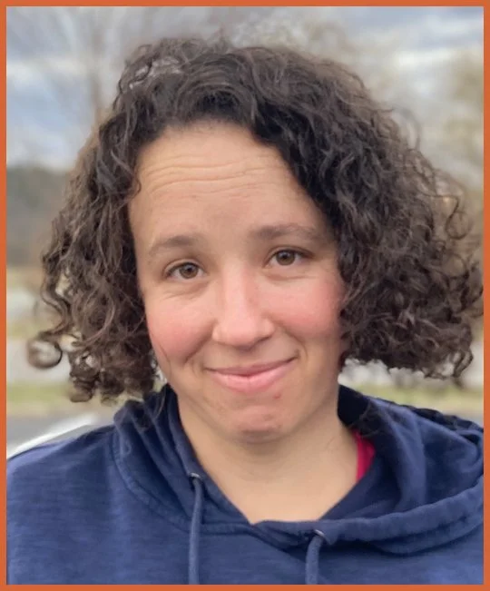 A woman with short, curly brown hair wearing a navy hoodie outdoors.