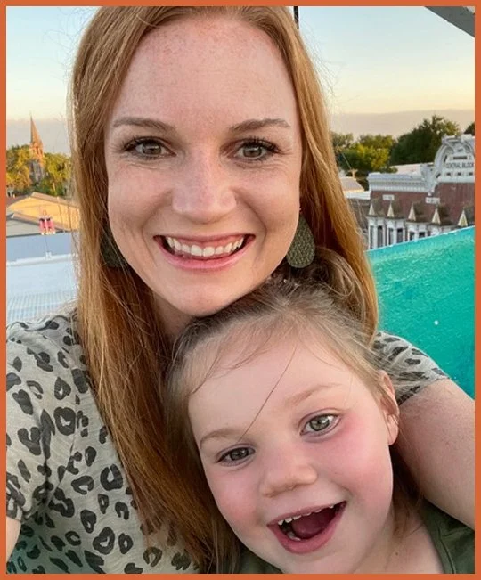 A woman with long red hair and an off-shoulder leopard print top smiling with a young girl with light brown hair, blue eyes, and a wide open-mouthed smile.