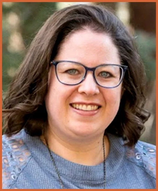 A woman with shoulder-length brown hair, glasses, and a blue sweater smiling outdoors with trees in the background.