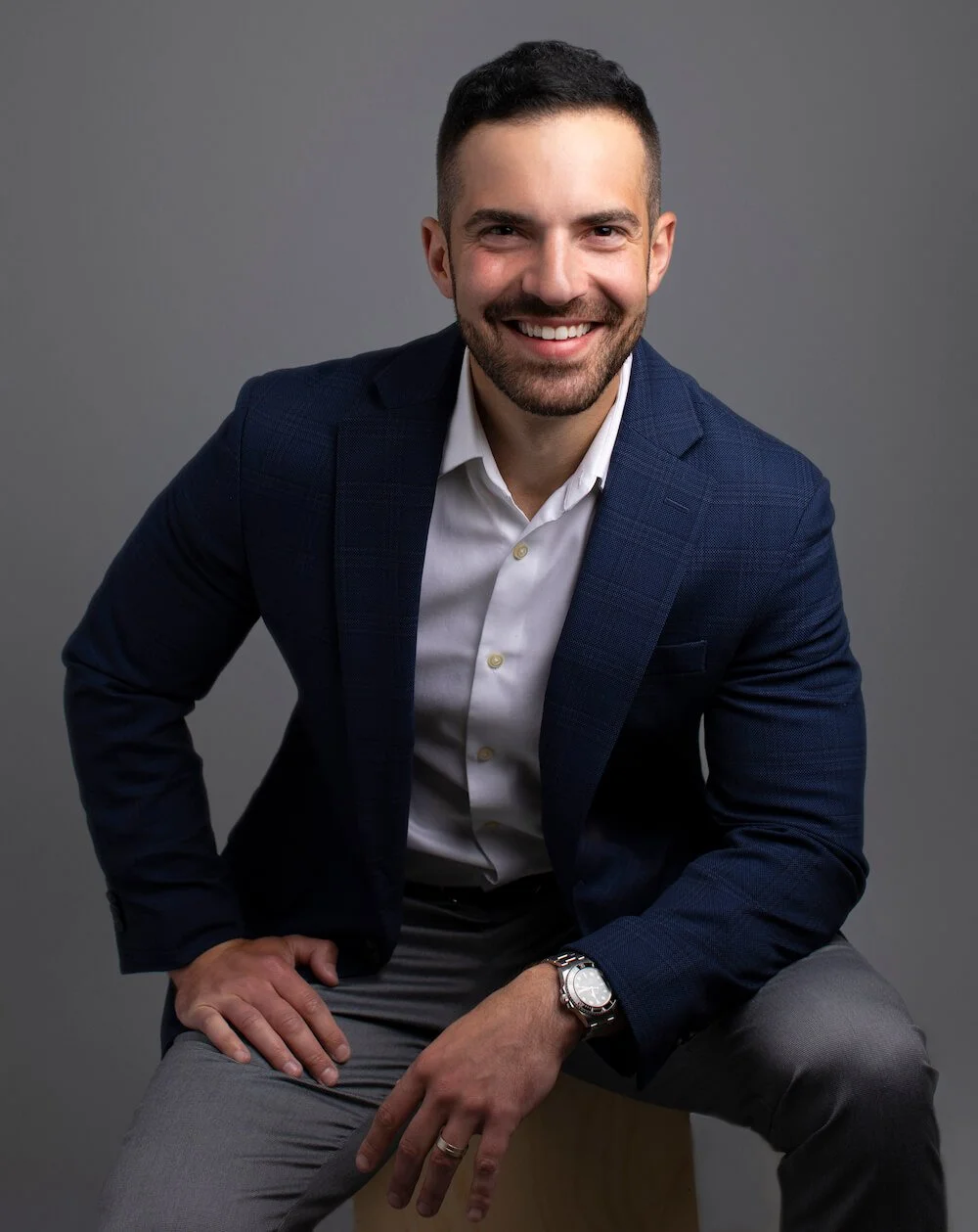 A young man with dark hair and a beard, smiling and wearing a dark blue blazer, white shirt, grey pants, sitting against a grey background.