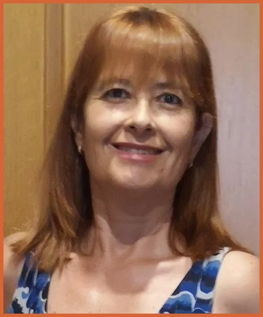 A smiling woman with shoulder-length red hair, wearing a blue patterned top, standing indoors with a wooden door in the background.
