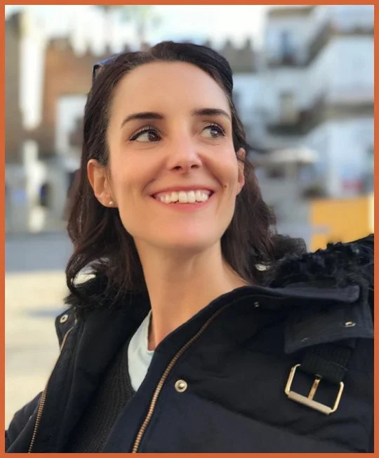 A woman smiling outdoors with buildings and a cloudy sky in the background, wearing a black jacket and a white shirt.