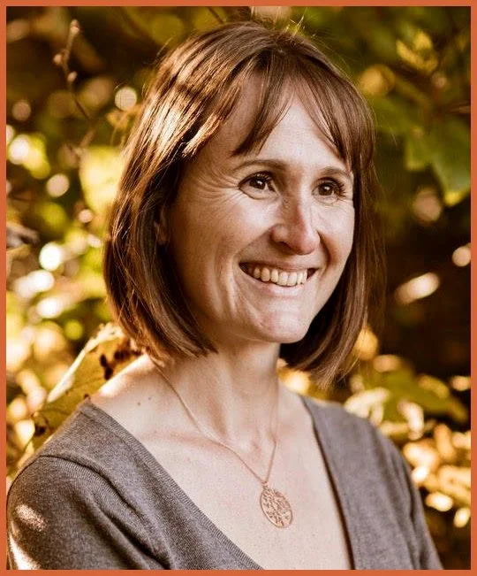A smiling woman with shoulder-length brown hair, wearing a gray top and a pendant necklace, outdoors with sunlight filtering through trees.