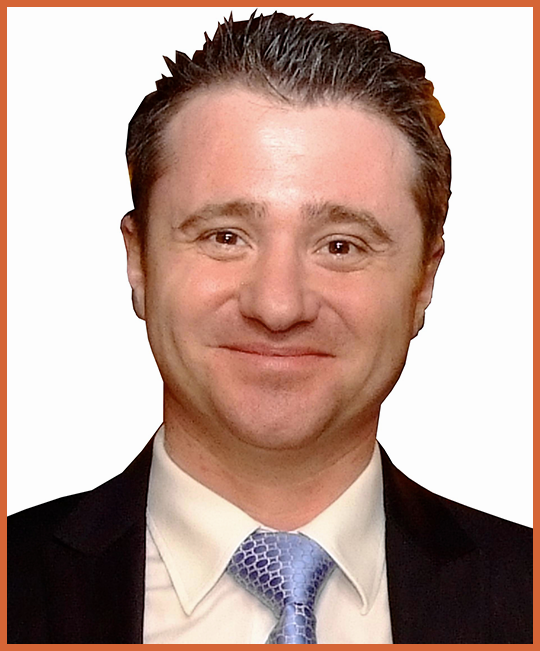 A professional man with short, dark, slicked-back hair wearing a dark suit, white shirt, and light blue tie, smiling at the camera.