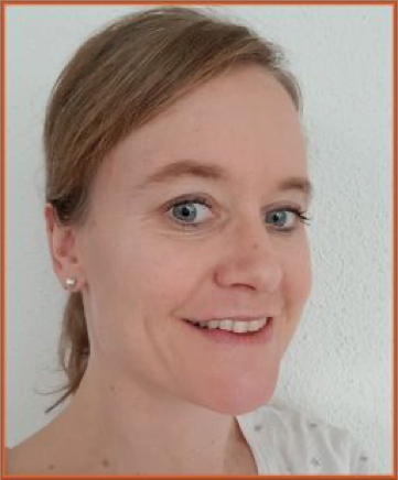 A woman with light skin and blue eyes smiling at the camera, with light brown hair pulled back, wearing small earrings and a white top, standing against a plain background.