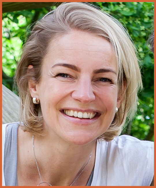 Close-up of a smiling woman with short blonde hair, wearing pearl earrings, a necklace, and a light-colored top, outdoors with green foliage in the background.