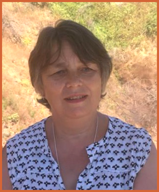 A woman with short brown hair standing outdoors, wearing a white and blue patterned top, with trees and a hill in the background.