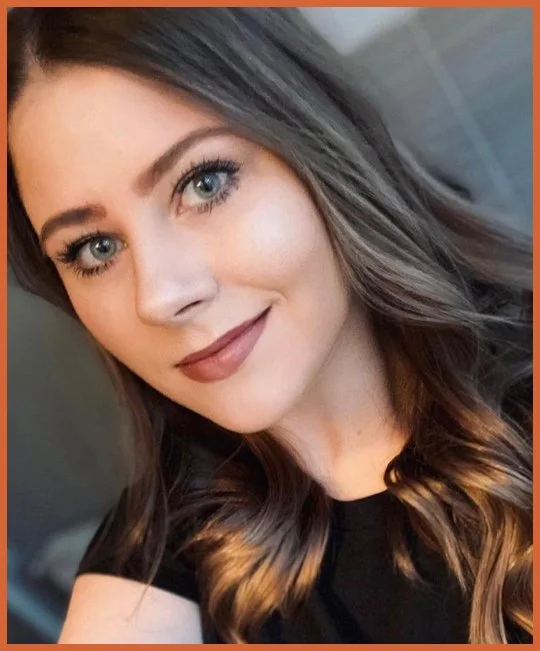 Close-up of a young woman with long wavy brown hair and blue eyes, smiling softly in a black top.