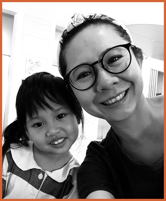 A woman with glasses and a headband smiling next to a young girl with dark hair and a striped shirt, both taking a selfie indoors.