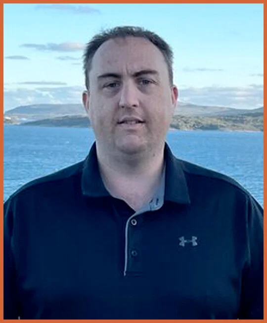 A man standing outdoors near a large body of water with hills in the background, wearing a dark polo shirt.