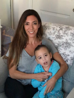 Nicole Zeitzer Johnson sitting on a couch, smiling, with her daughter Josie, in a blue shirt sitting on her lap, in a cozy living room.