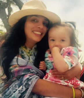 Nasha Fitter, co-founder of FOXG1 Research Foundation wearing a hat and a colorful top holding her daughter Amara who is wearing a floral dress outdoors.
