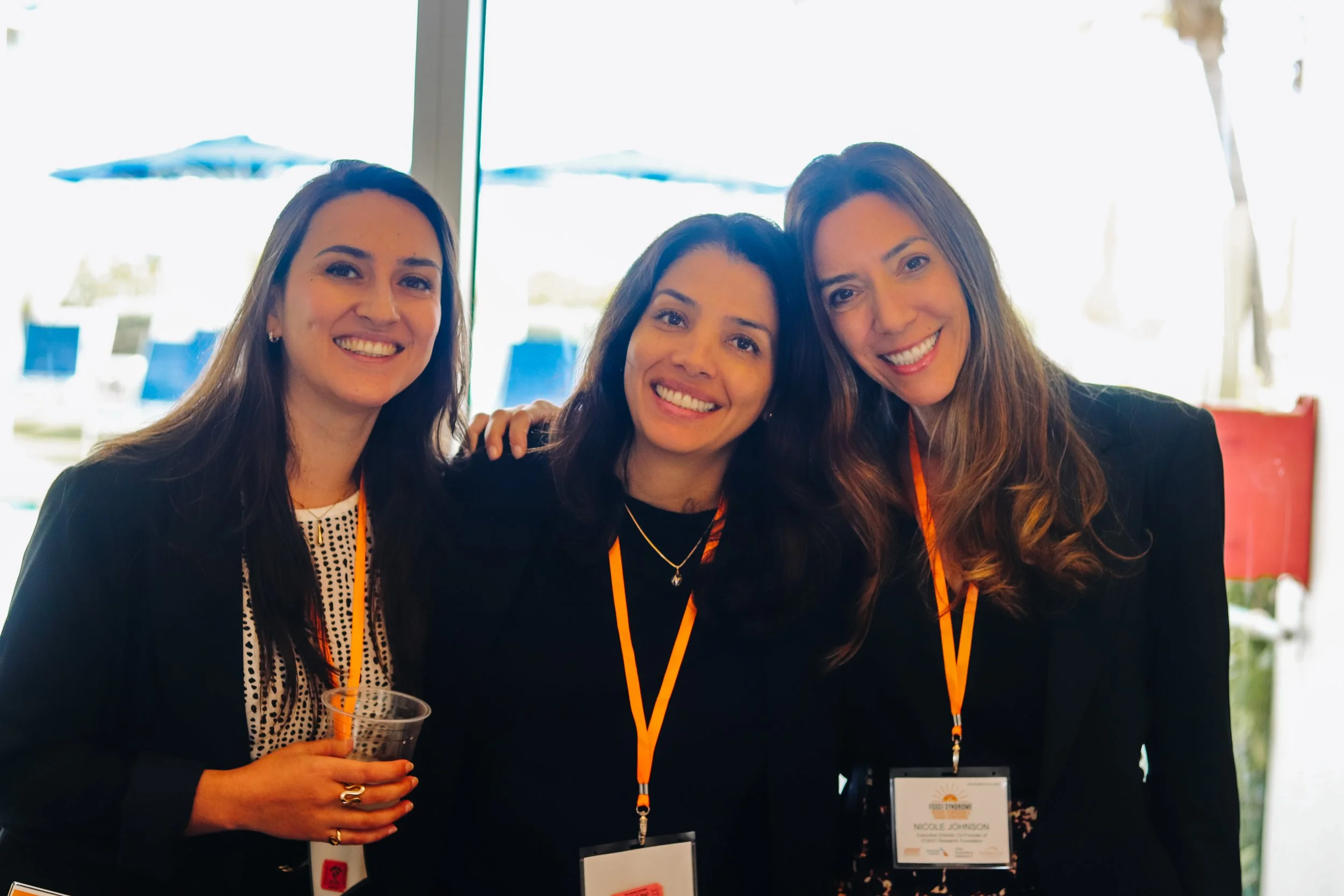 Three women smiling at a conference, wearing black blazers and orange lanyards.