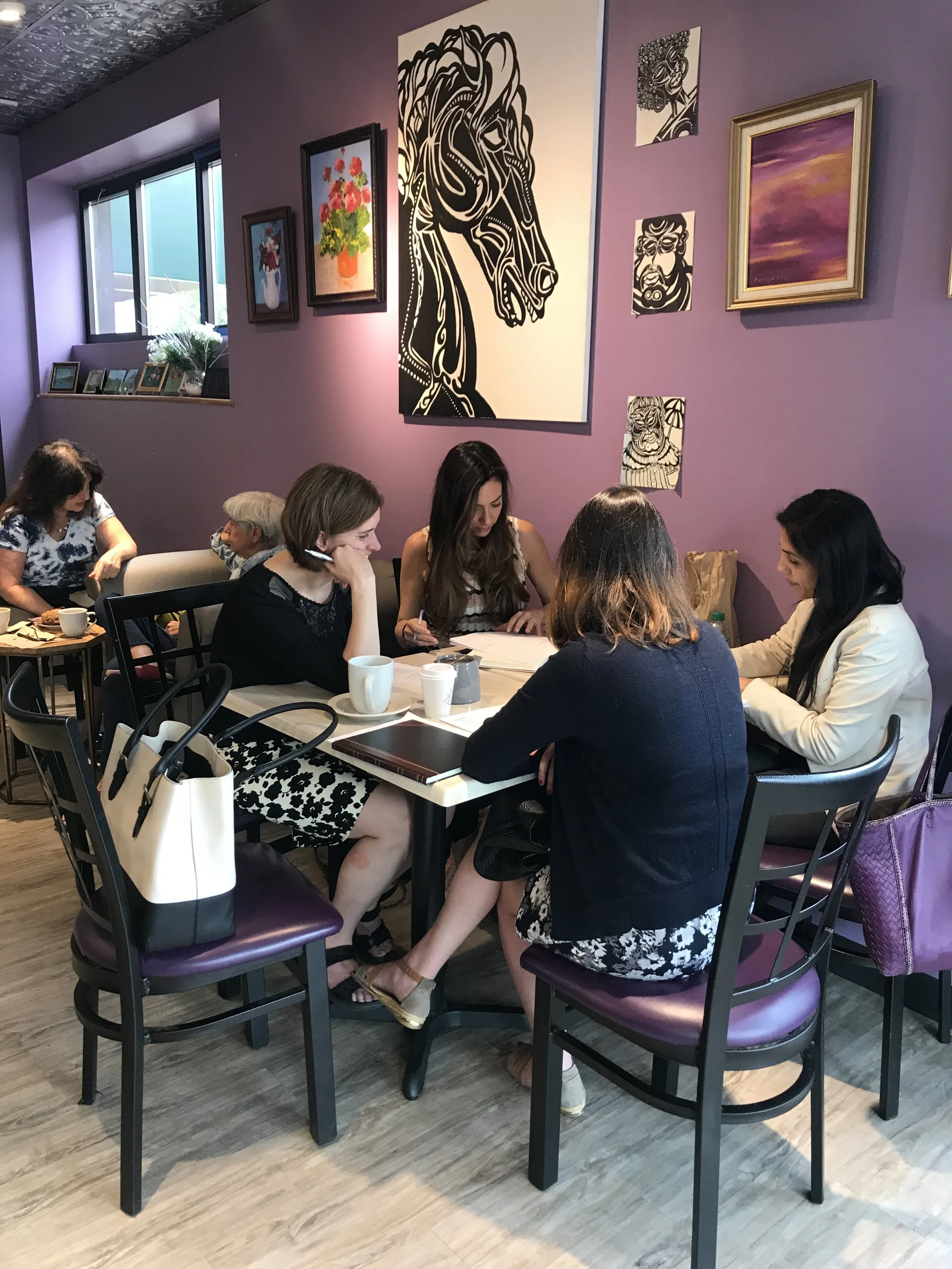 A group of five women sitting around a table in a cafe, engaged in a discussion or working together. The cafe has purple walls decorated with various framed paintings and artwork.