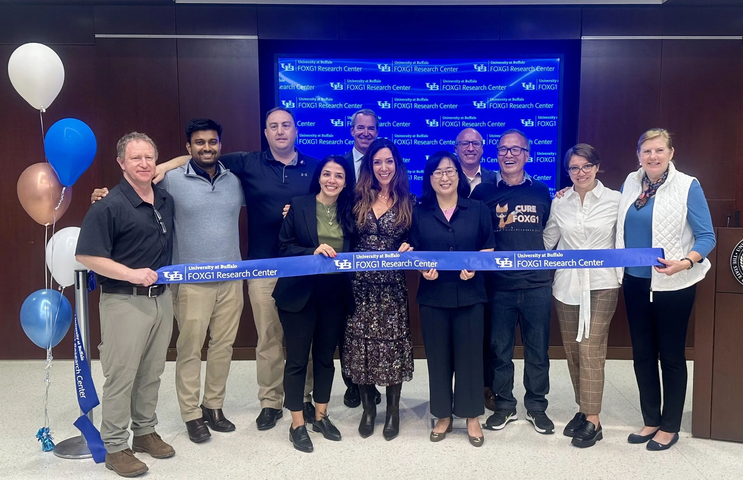 A group of people standing together in front of a blue informational backdrop for the FOXG1 Research Center at University at Buffalo. They are holding a blue ribbon that reads 'University at Buffalo FOXG1 Research Center' and smiling for the photo. There are balloons on the left side.