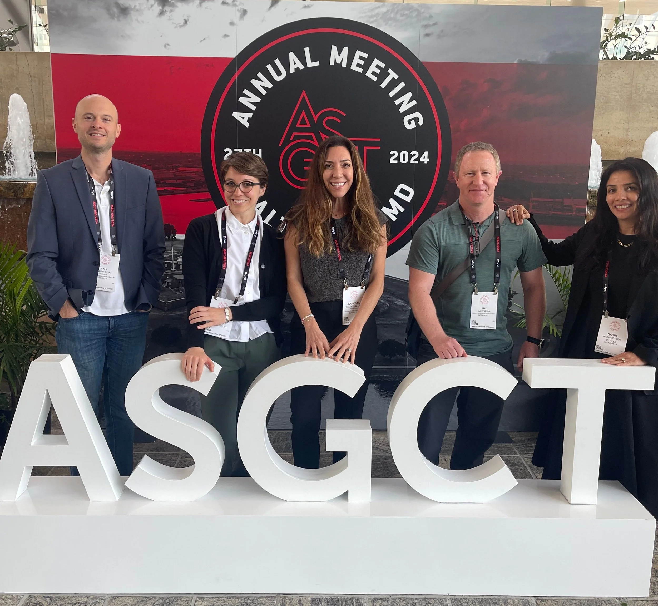 Group of five people standing behind large white letters spelling ASGCT at an Annual Meeting event in 2024 in Maryland, with a backdrop displaying event details and a sunset sky.