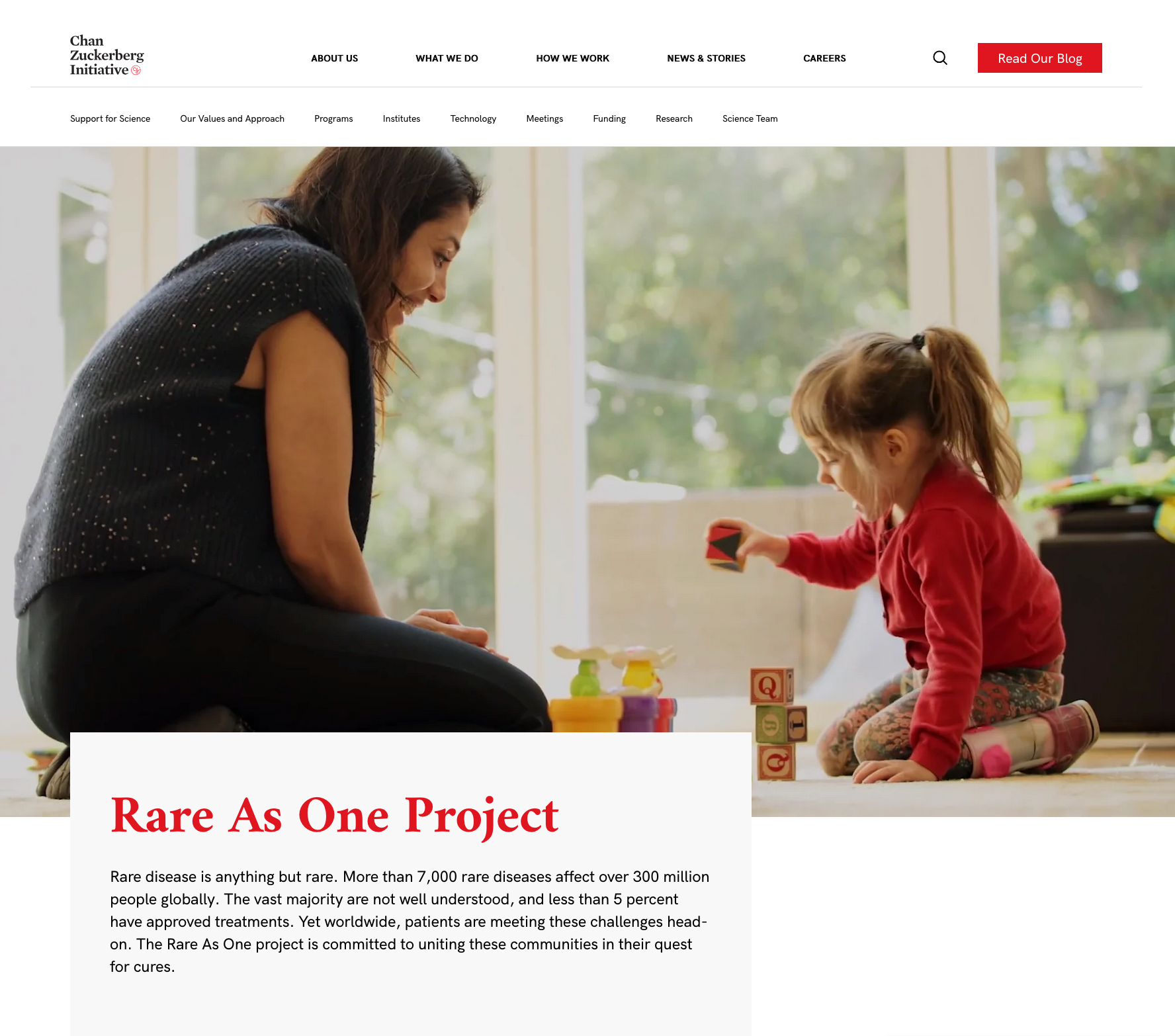 A woman and a young girl playing with blocks and toys indoors near large windows with greenery outside.