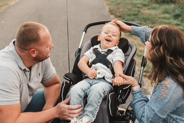 A young boy with cancer sitting in a stroller, laughing and holding hands with a man and a woman outdoors, with the woman gently touching his head.
