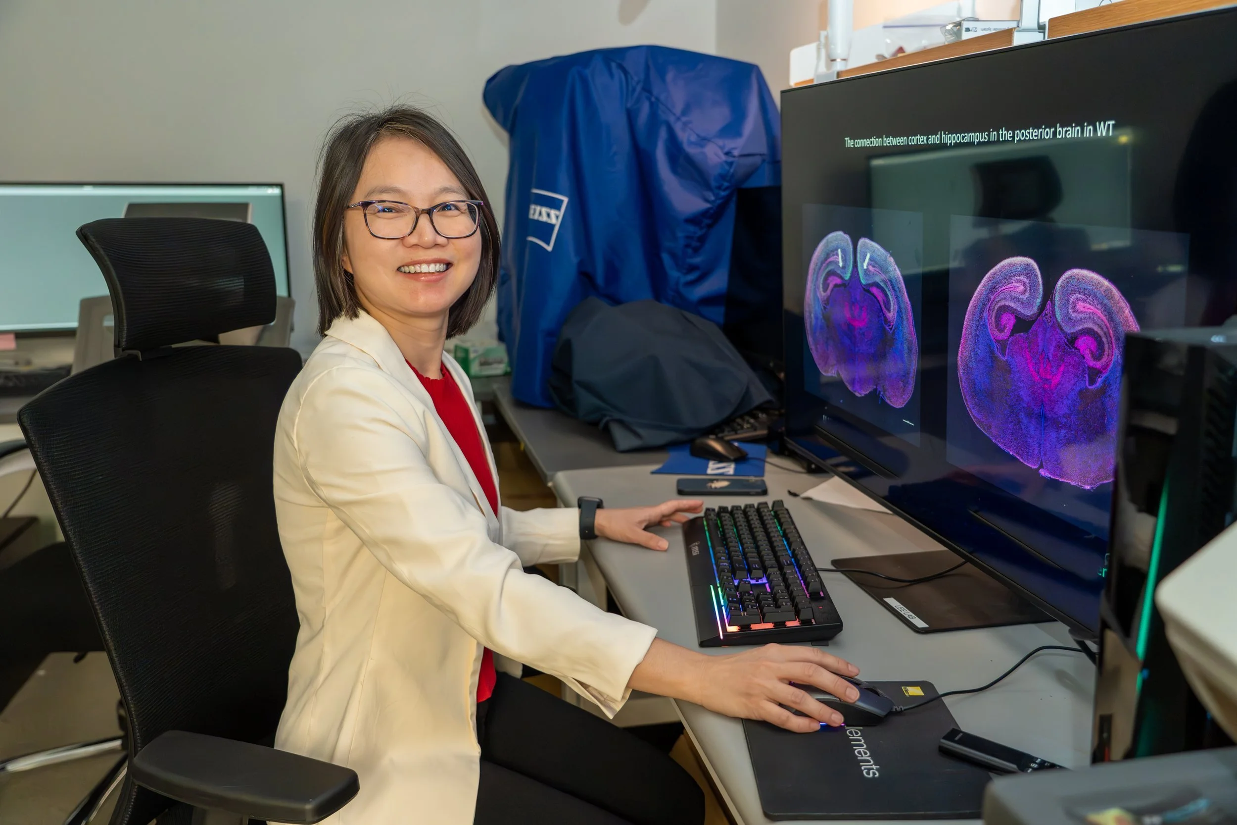 A woman with glasses is sitting at a desk in a workspace, smiling at the camera. She is operating a computer with a large monitor displaying brain scan images. The workspace has multiple computer screens and equipment, with a blue cover or bag in the background.