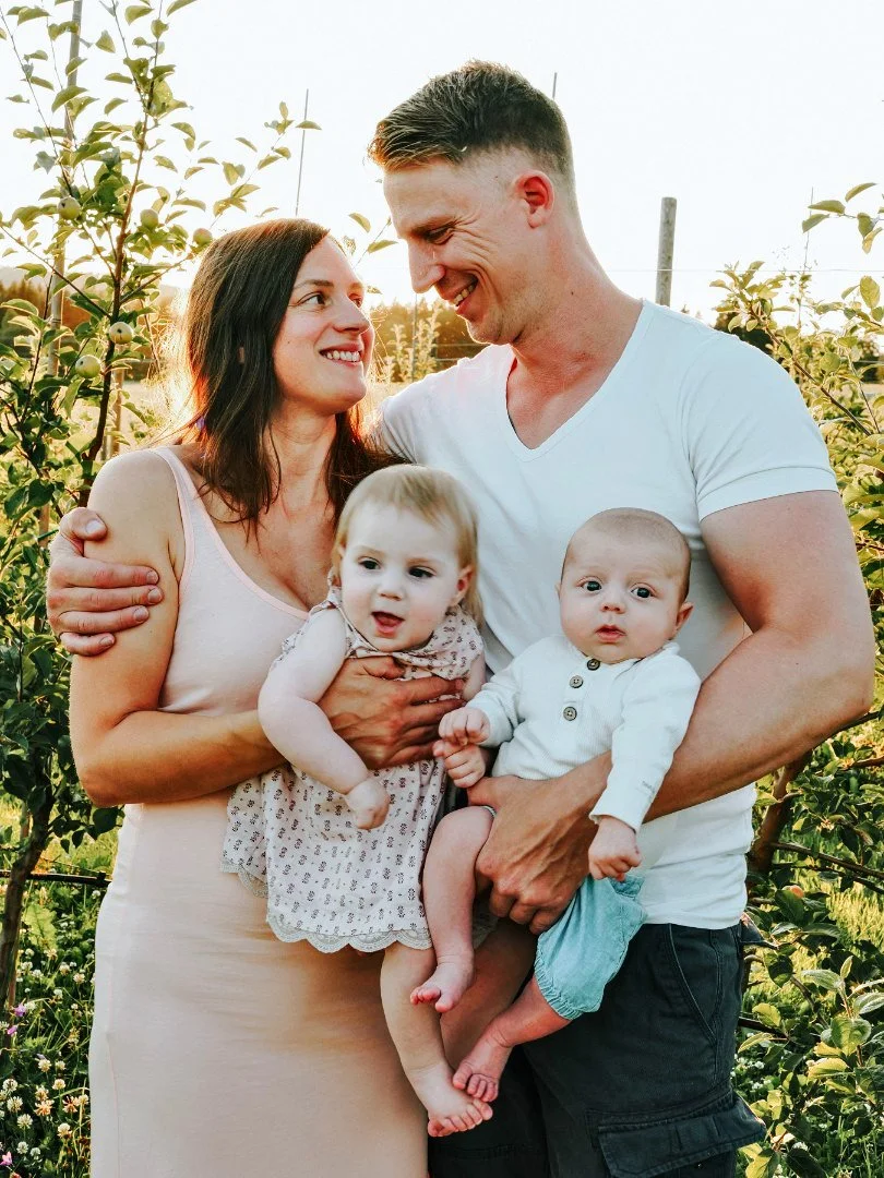 A happy family of four standing outdoors in a garden during sunset. The mother and father are smiling and looking at each other, holding their two young children, a girl and a boy, who are also looking at the camera.