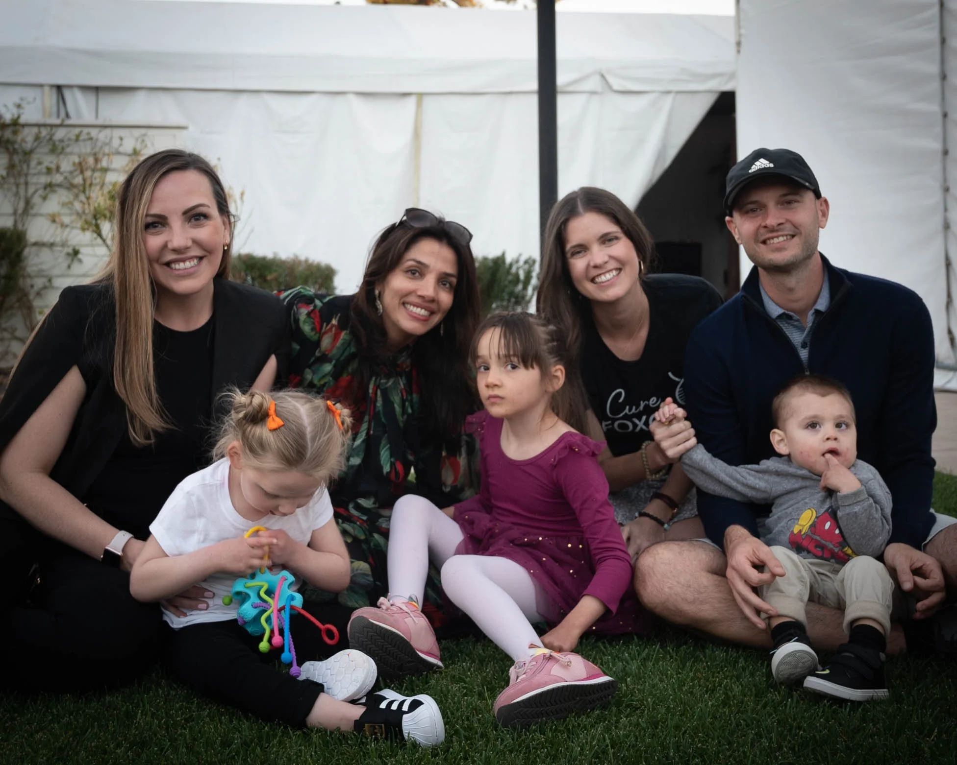Family and friends sitting on grass at an outdoor gathering, smiling for the camera, with a white tent in the background.