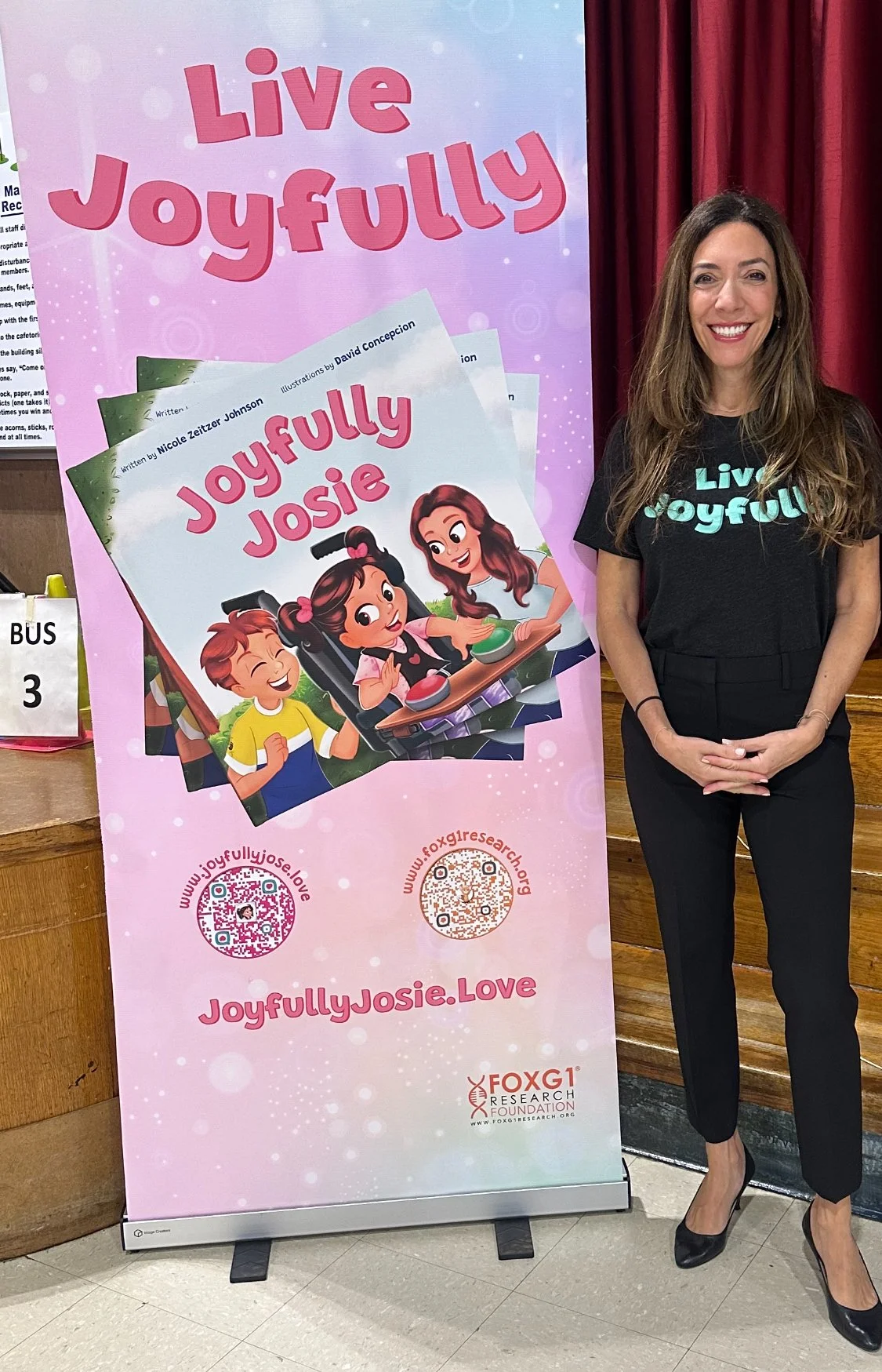 A woman standing next to a large pink promotional poster for the book 'Joyfully Josie' by Nicole Zeitzer Johnson. The poster features animated children playing and the book's title and website information.