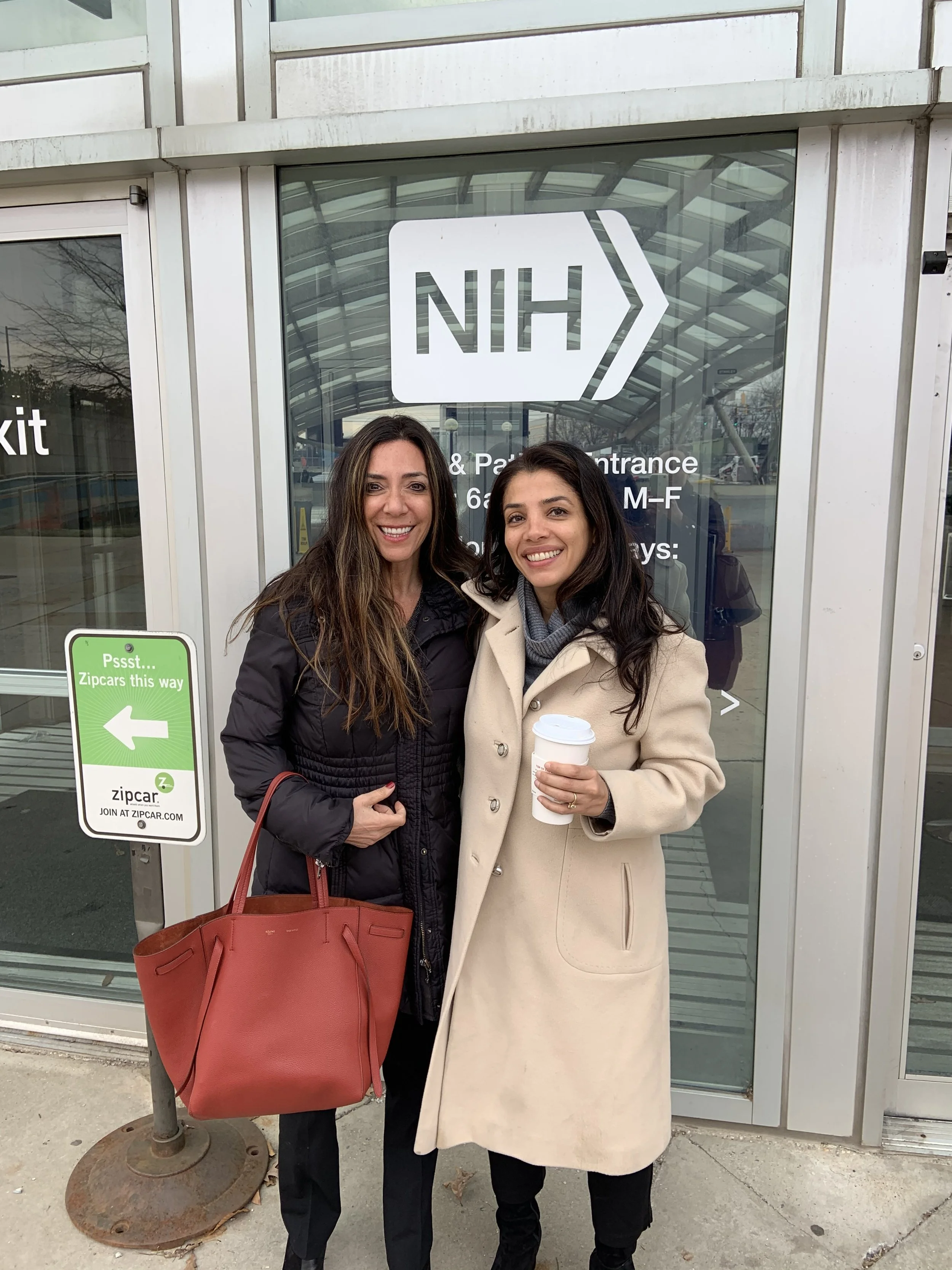 Two women standing outside of the NIH building with a glass entrance, smiling at the camera. One woman is wearing a black jacket and carrying a large red handbag. The other woman is wearing a beige coat and holding a coffee cup.