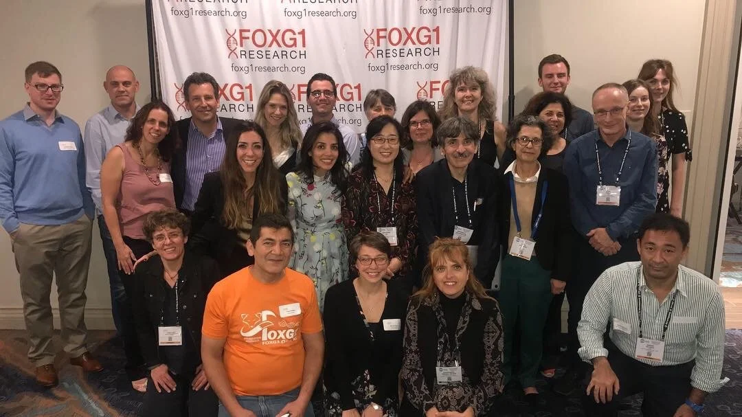 Group photo of diverse professionals at a conference, standing in front of a backdrop with the FOXG1 research logo.