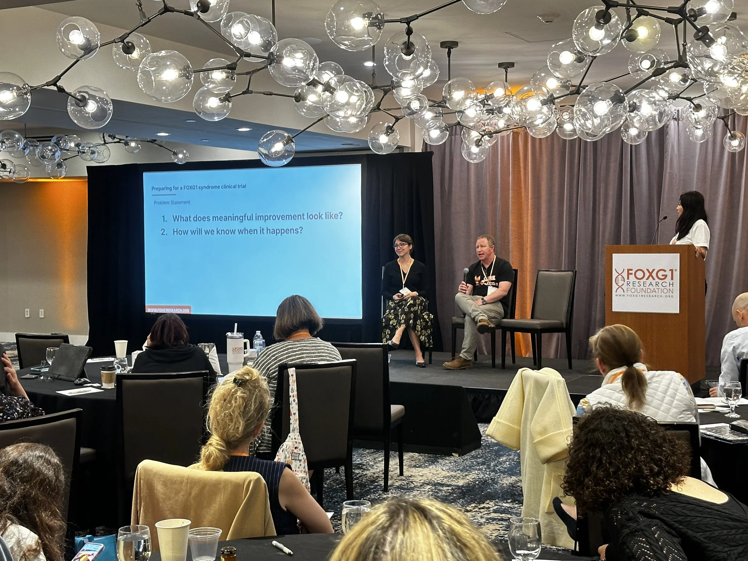 A conference room with a stage and a presentation screen showing two questions related to clinical trials. There are three speakers on the stage, one woman standing at a podium labeled FoxG1 Research Foundation, a woman seated on the stage, and a man