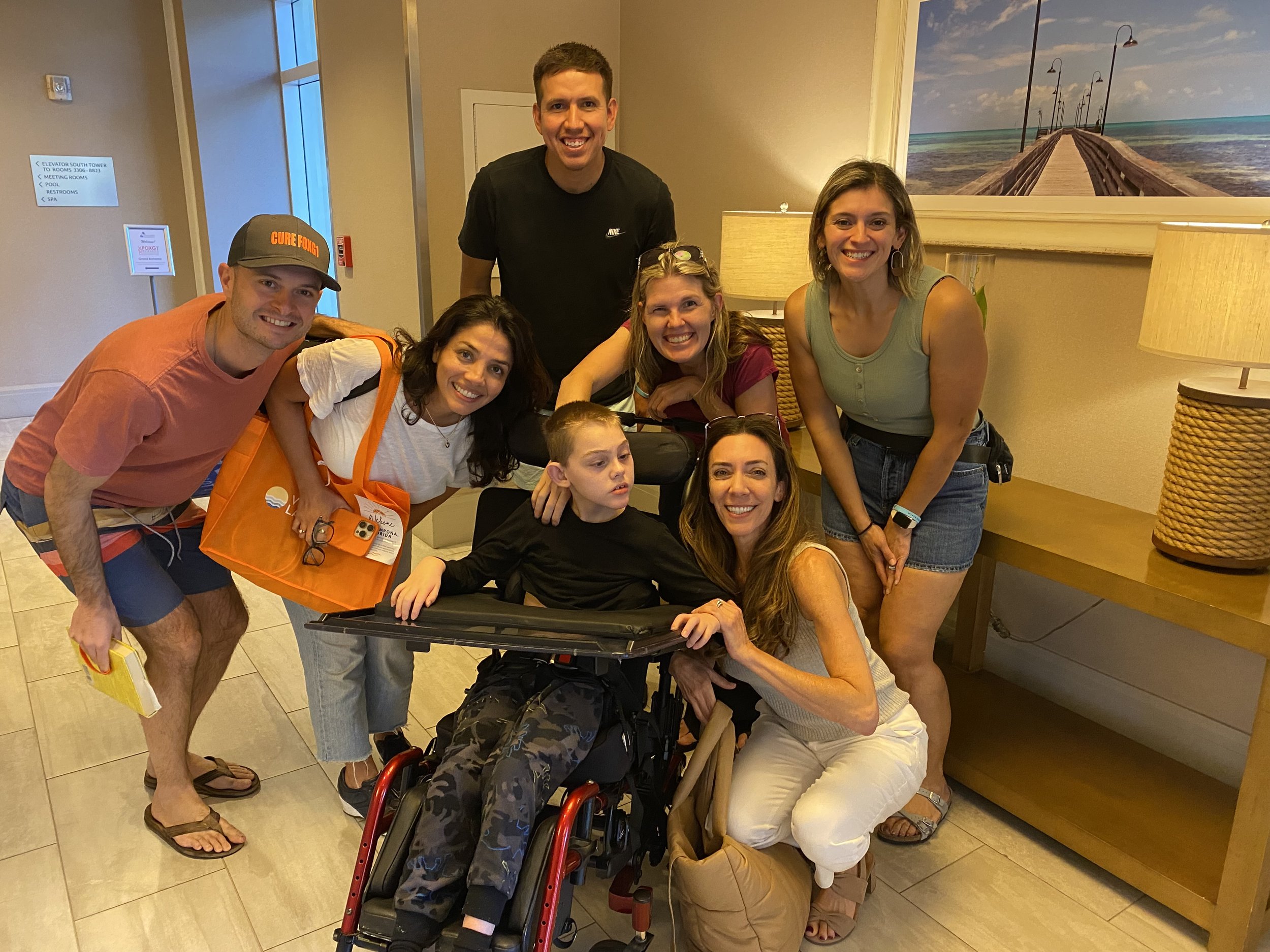 Group of seven people, including a young boy in a wheelchair, smiling inside a hotel lobby.