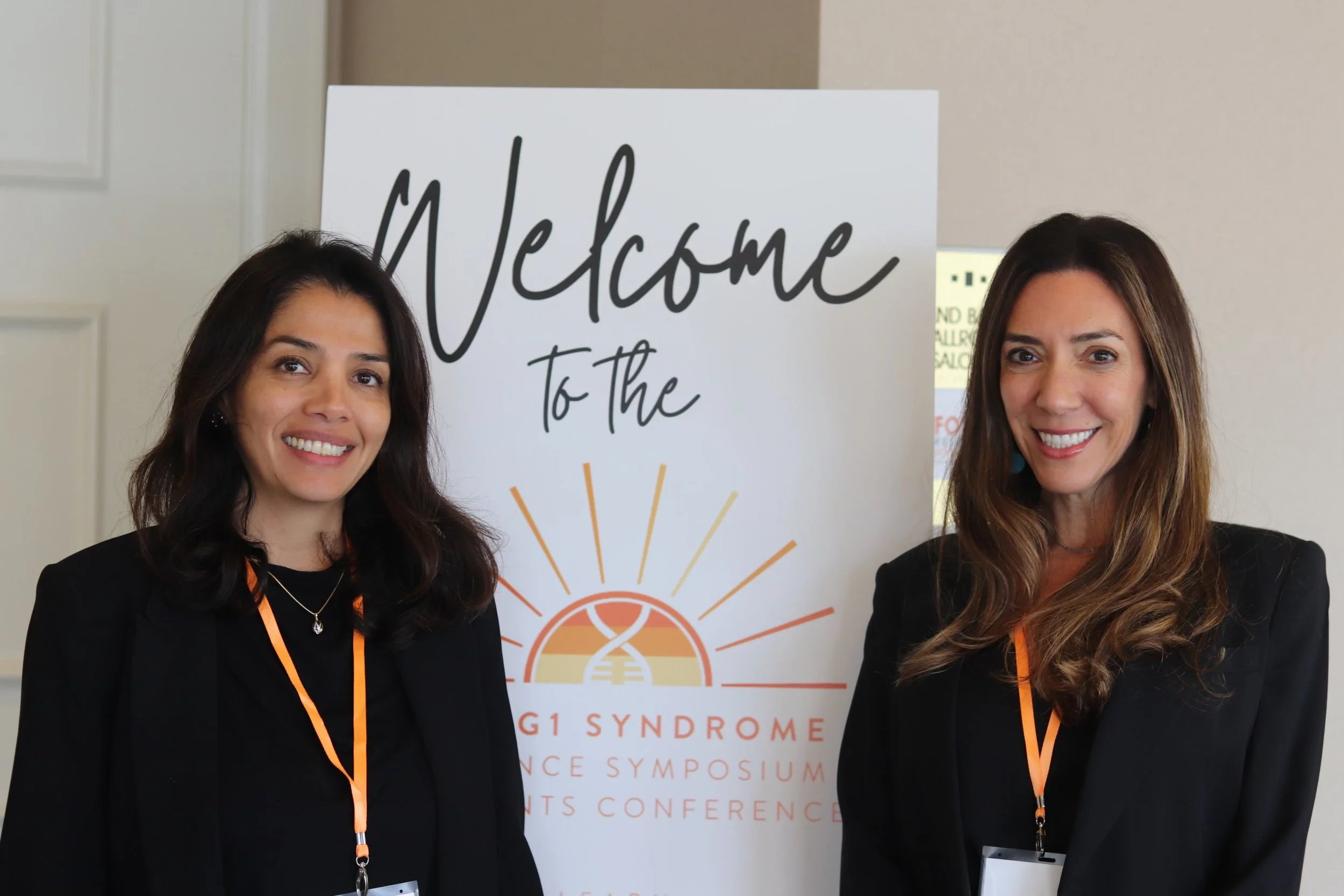 Two smiling women standing in front of a welcome sign at a FOXG1 Science conference, with lanyards and badges.