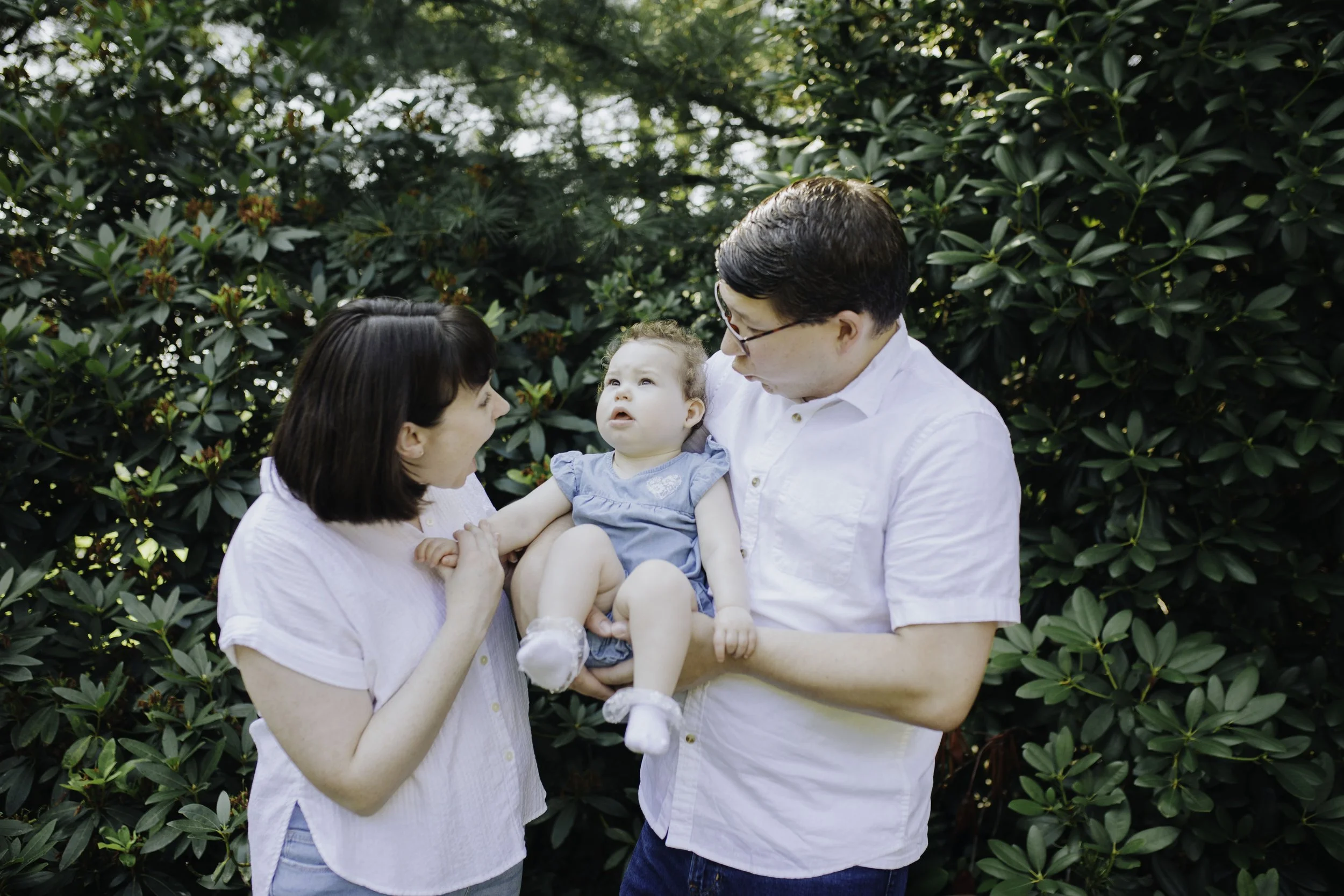 A family of three, a man, a woman, and a young girl, standing outdoors in front of green bushes. The man is holding the girl, who is looking at her mother. The woman appears to be upset or crying.